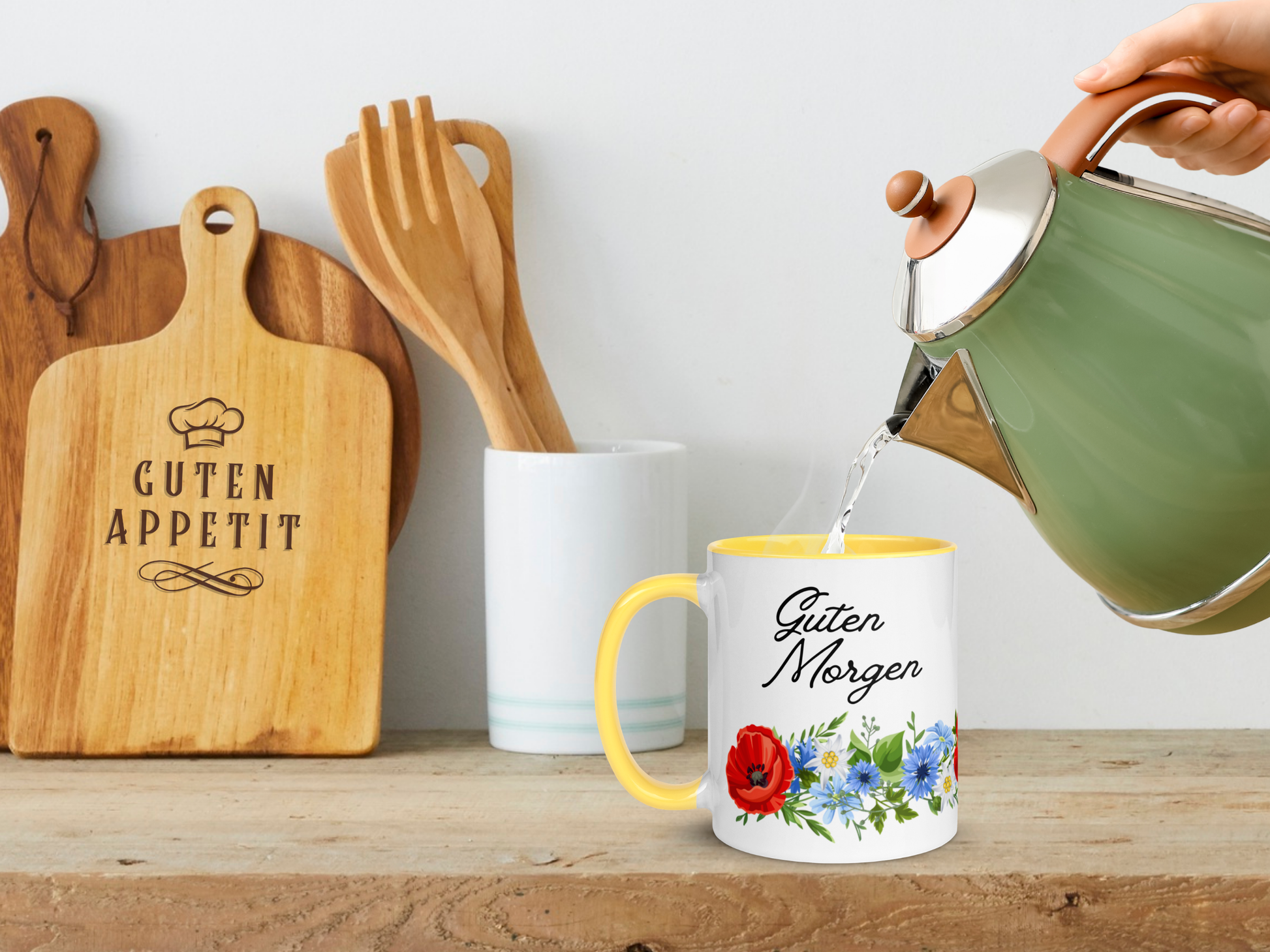 Person pouring hot water into a mug with floral design and 'Guten Morgen' text, surrounded by kitchen items.