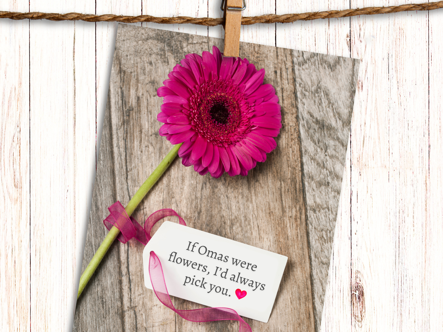 Greeting card featuring a pink Gerbera daisy against a wood background with a  tag, attached with pink ribbon, that says "If Omas were Flowers, I'd always pick you" and heart emoji.